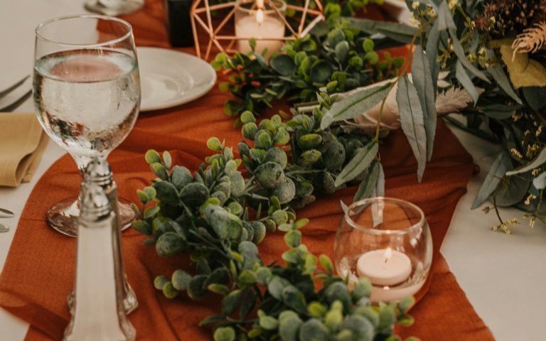 Low greenery and glass candle holders arranged along a rust-colored dining table runner