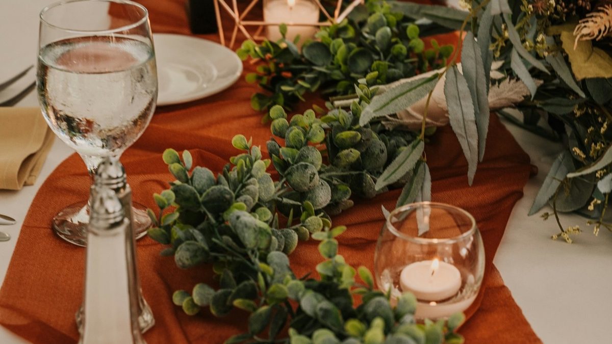 Greenery and glass candle holders arranged along a rust-colored dining table runner