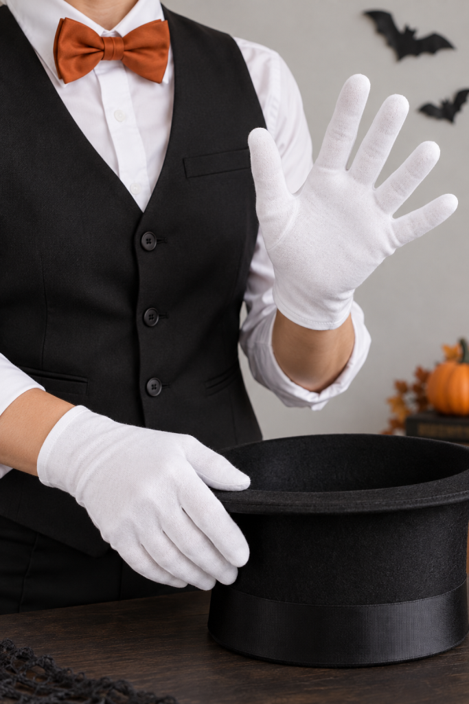An adult wearing a black waistcoat, white shirt, and rust bow tie raises one white-gloved palm while resting the other gloved hand beside a black top hat.